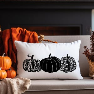 Könnte beinhalten: Ein weißes rechteckiges Kissen mit drei schwarzen Kürbismotiven. Der mittlere Kürbis ist einfarbig schwarz, während die anderen beiden florale Muster aufweisen. Das Kissen liegt auf einem beigen Sofa, mit orangefarbenen Kürbissen und Herbstdekoration im Hintergrund.