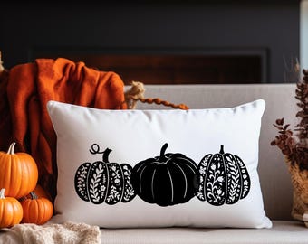 Puede incluir: Un cojín rectangular blanco con tres diseños de calabazas negras. La calabaza central es negra lisa, mientras que las otras dos tienen diseños florales. El cojín está sobre un sofá beige, con calabazas naranjas y decoración otoñal en el fondo.
