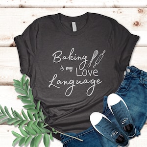 May include: A dark gray t-shirt with the text "Baking is my Love Language" in white lettering. The text is accompanied by a white whisk and rolling pin graphic.