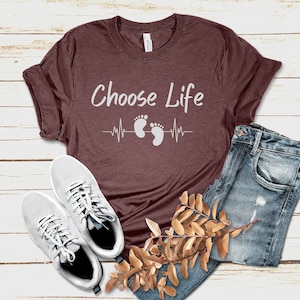 Puede incluir: Una camiseta marrón con el texto "Choose Life" y un gráfico de latido del corazón con dos huellas de pies. La camiseta está sobre una superficie de madera blanca con un par de zapatillas blancas y un par de pantalones vaqueros azules.