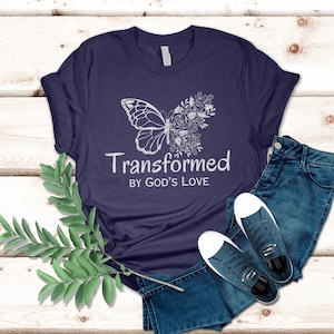 May include: A navy blue t-shirt featuring a white butterfly and floral design. The text "Transformed by God's Love" is printed on the shirt.