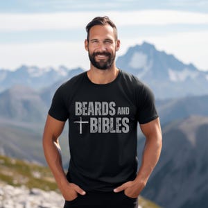 May include: Black t-shirt featuring the text "BEARDS AND BIBLES" in distressed white lettering, with a cross symbol. The shirt is worn by a person with a beard, set against a mountain backdrop.