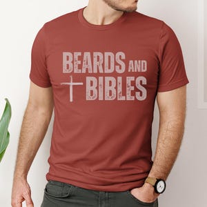 May include: A rust-coloured t-shirt featuring the text "BEARDS and BIBLES" in a faded, light grey font. A white cross is positioned below "BEARDS". The shirt has a crew neck and is being worn.