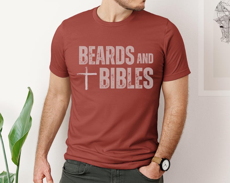 May include: A rust-coloured t-shirt featuring the text "BEARDS and BIBLES" in a faded, light grey font. A white cross is positioned below "BEARDS". The shirt has a crew neck and is being worn.