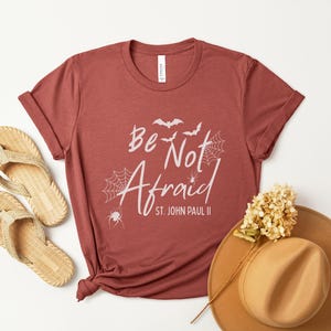 May include: A maroon t-shirt with the words "Be Not Afraid" and "St. John Paul II" printed in white, along with spiderwebs, bats, and a spider graphic. The shirt is styled with a straw hat and sandals.