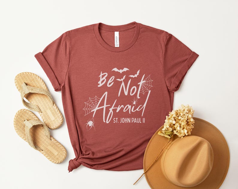 May include: A maroon t-shirt with the words "Be Not Afraid" and "St. John Paul II" printed in white, along with spiderwebs, bats, and a spider graphic. The shirt is styled with a straw hat and sandals.