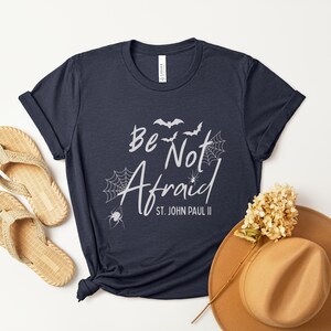 May include: Navy blue t-shirt with the text "Be Not Afraid" and "St. John Paul II" in white script. The design includes bats, spiderwebs, and spiders. A pair of woven sandals and a brown hat are also in the image.