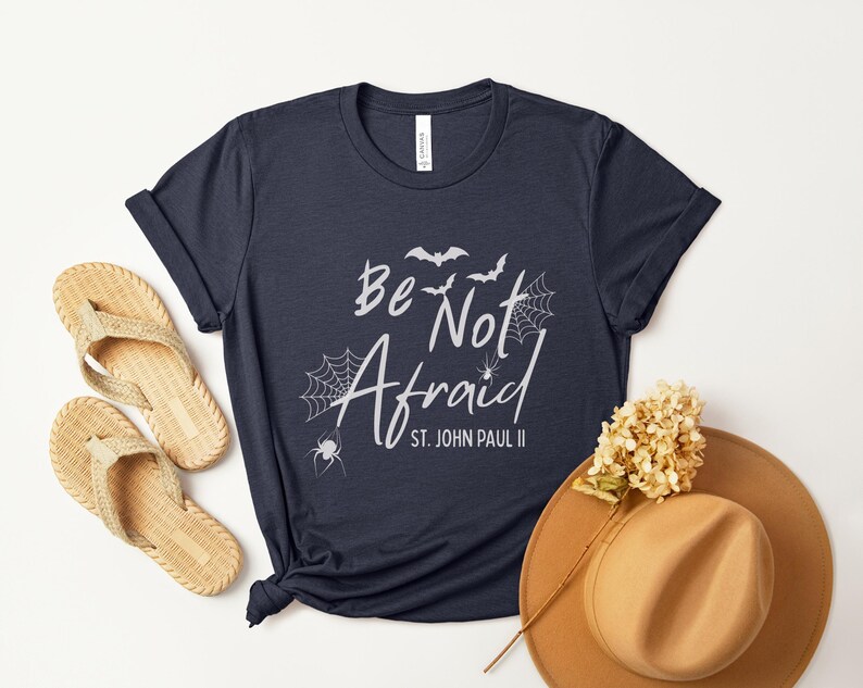 May include: Navy blue t-shirt with the text "Be Not Afraid" and "St. John Paul II" in white script. The design includes bats, spiderwebs, and spiders. A pair of woven sandals and a brown hat are also in the image.