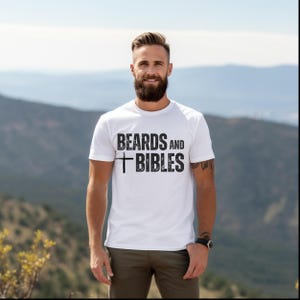 May include: White t-shirt featuring the text "BEARDS AND BIBLES" in black, with a cross symbol. The person wearing the shirt has a beard and is standing outside with a mountain range in the background.
