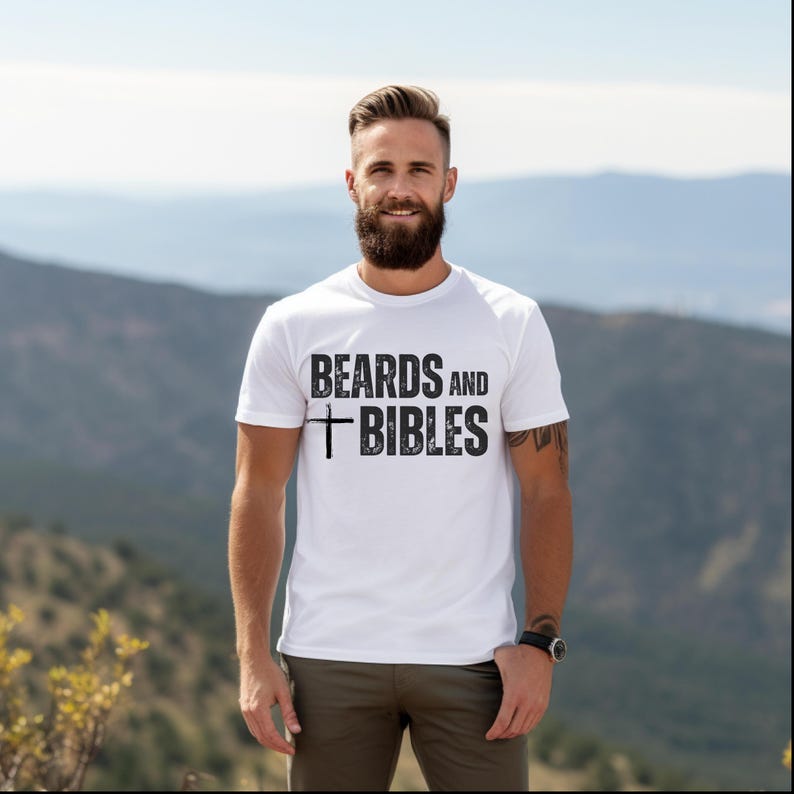 May include: White t-shirt featuring the text "BEARDS AND BIBLES" in black, with a cross symbol. The person wearing the shirt has a beard and is standing outside with a mountain range in the background.