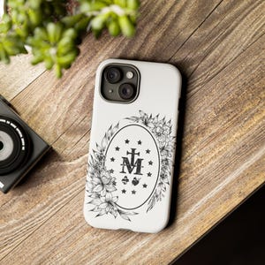 K&ouml;nnte beinhalten: Wei&szlig;e iPhone-H&uuml;lle mit einer schwarz-wei&szlig;en Illustration eines Blumenkranzes, der die Wundermedaille umgibt, die die Jungfrau Maria, ein Kreuz und die Buchstaben "M" und "A" zeigt.