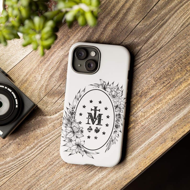 K&ouml;nnte beinhalten: Wei&szlig;e iPhone-H&uuml;lle mit einer schwarz-wei&szlig;en Illustration eines Blumenkranzes, der die Wundermedaille umgibt, die die Jungfrau Maria, ein Kreuz und die Buchstaben "M" und "A" zeigt.