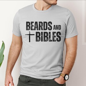 May include: A light grey t-shirt featuring the text "BEARDS AND BIBLES" in distressed black font, with a black cross graphic. The shirt has short sleeves and a crew neck.