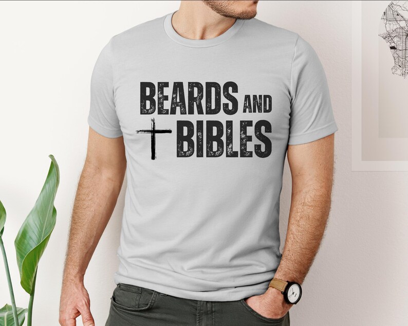 May include: A light grey t-shirt featuring the text "BEARDS AND BIBLES" in distressed black font, with a black cross graphic. The shirt has short sleeves and a crew neck.