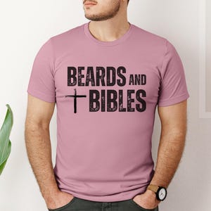 May include: A short-sleeved, dusty rose t-shirt featuring the black text "BEARDS and BIBLES" and a cross. The shirt is made of a soft material. The text and cross are printed in a distressed style.