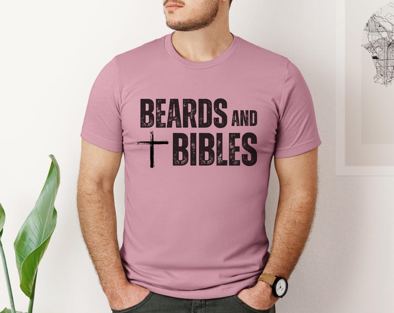 May include: A short-sleeved, dusty rose t-shirt featuring the black text "BEARDS and BIBLES" and a cross. The shirt is made of a soft material. The text and cross are printed in a distressed style.