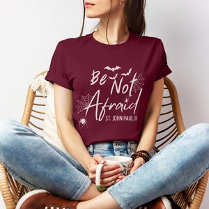 May include: A maroon t-shirt with the words "Be Not Afraid" and "St. John Paul II" in white script. The design includes bats, spiderwebs, and a spider. The person is wearing blue jeans and brown shoes.