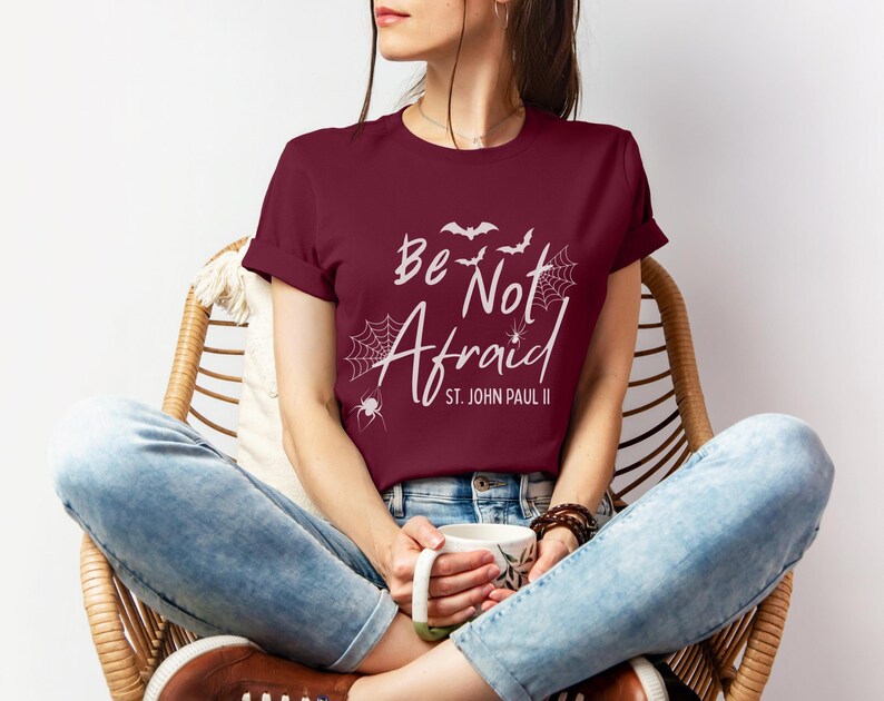 May include: A maroon t-shirt with the words "Be Not Afraid" and "St. John Paul II" in white script. The design includes bats, spiderwebs, and a spider. The person is wearing blue jeans and brown shoes.