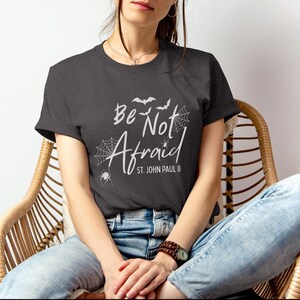 May include: Dark gray t-shirt with the words "Be Not Afraid" and "St. John Paul II" in white script. The design includes bats, spiderwebs, and a spider. The shirt is worn by a person sitting in a wicker chair.