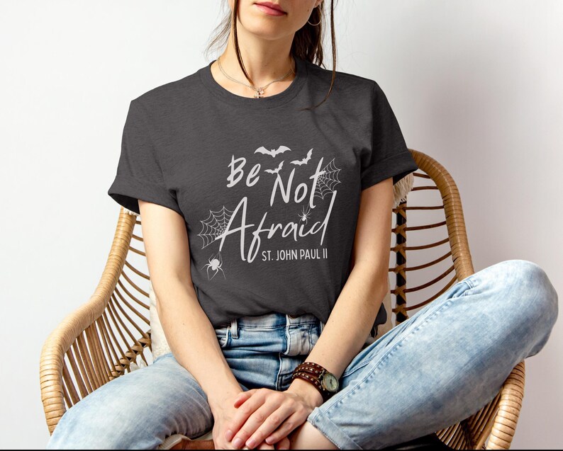 May include: Dark gray t-shirt with the words "Be Not Afraid" and "St. John Paul II" in white script. The design includes bats, spiderwebs, and a spider. The shirt is worn by a person sitting in a wicker chair.