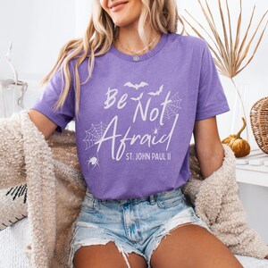 May include: A purple t-shirt with the words "Be Not Afraid" in white, along with spiderwebs, bats, and the text "St. John Paul II." The shirt is worn with denim shorts and a cream-colored sweater. A decorative pumpkin and basket are in the background.