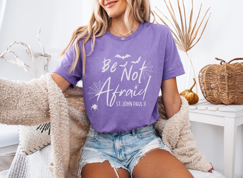 May include: A purple t-shirt with the words "Be Not Afraid" in white, along with spiderwebs, bats, and the text "St. John Paul II." The shirt is worn with denim shorts and a cream-colored sweater. A decorative pumpkin and basket are in the background.