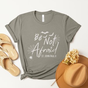 May include: A sage green t-shirt with the words "Be Not Afraid" and a spiderweb and bat graphic. The shirt is styled with woven sandals and a brown hat with a flower.