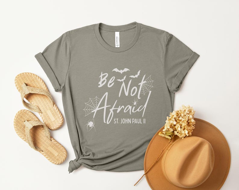 May include: A sage green t-shirt with the words "Be Not Afraid" and a spiderweb and bat graphic. The shirt is styled with woven sandals and a brown hat with a flower.