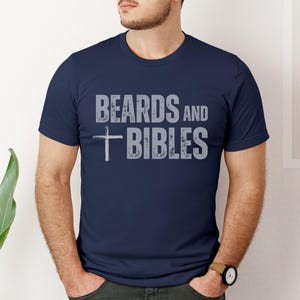 May include: A navy blue crew neck t-shirt featuring the text "BEARDS AND BIBLES" in grey, with a cross symbol. The t-shirt is being worn by a person.