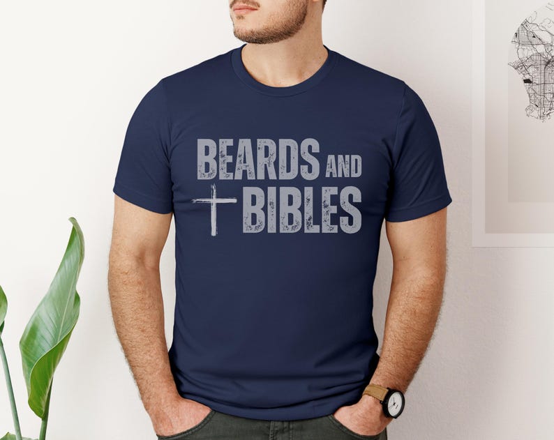 May include: A navy blue crew neck t-shirt featuring the text "BEARDS AND BIBLES" in grey, with a cross symbol. The t-shirt is being worn by a person.