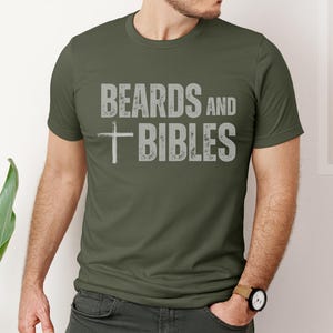 May include: An olive green t-shirt featuring the text "BEARDS and BIBLES" in distressed grey lettering and a grey cross graphic. The t-shirt has a crew neck design.