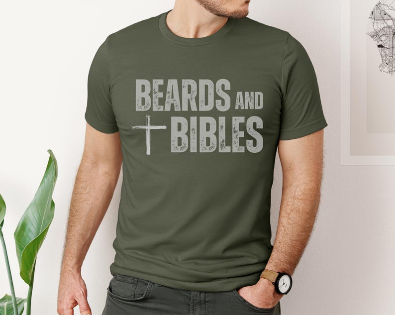 May include: An olive green t-shirt featuring the text "BEARDS and BIBLES" in distressed grey lettering and a grey cross graphic. The t-shirt has a crew neck design.