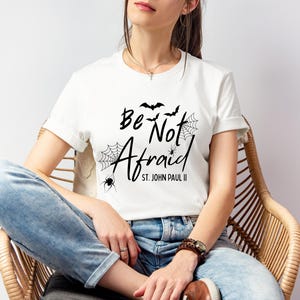 May include: White t-shirt with the phrase "Be Not Afraid" in black script, accompanied by bat and spiderweb graphics. The text "St. John Paul II" is below the main text. The shirt is worn by a person.
