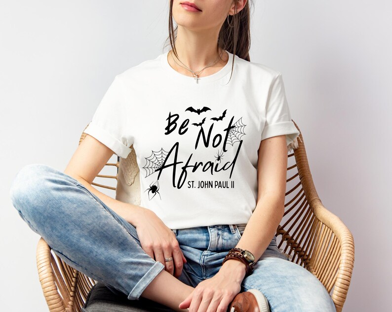 May include: White t-shirt with the phrase "Be Not Afraid" in black script, accompanied by bat and spiderweb graphics. The text "St. John Paul II" is below the main text. The shirt is worn by a person.