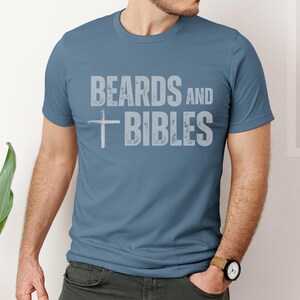 May include: A blue crew neck t-shirt with the text "BEARDS and BIBLES" in a distressed, light grey font. A white cross is centered below the word "BEARDS". The shirt is made of a soft material.