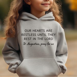 Op de afbeelding: Een grijs hoodie met de tekst "OUR HEARTS ARE RESTLESS UNTIL THEY REST IN THE LORD St. Augustine, pray for us" in zwart gedrukt.