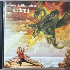 May include: CD case for the album "Trilogy" by Yngwie J. Malmsteen. The cover art features a three-headed dragon breathing fire at a person holding a guitar. The background is a fiery sky with rocky terrain.