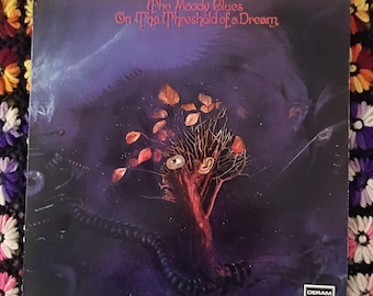 May include: A square vinyl record cover for The Moody Blues album On The Threshold of a Dream. The artwork features a dark purple, cosmic background with a central tree-like structure, human ears, and orange leaves, with Deram London text.