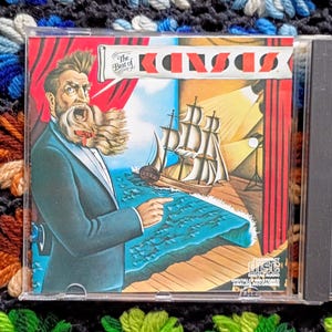 May include: A CD case featuring the album "The Best of Kansas." The cover art depicts a man in a suit with a flowing beard, a ship, and the band's name in red and white. The CD case is clear plastic.