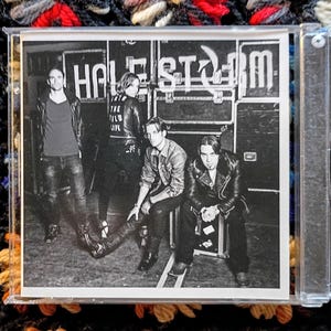 May include: Black and white CD cover featuring the band Halestorm. The band members are posed in front of equipment cases. One member has "THE WILD LIFE" printed on the back of their jacket.