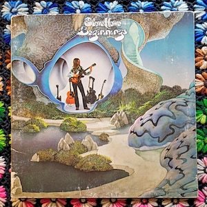 May include: Album cover for Steve Howe's "Beginnings." The artwork features a musician playing a guitar within a blue, cloud-like frame. Other guitars are visible. The background has a landscape with water, rocks, and vegetation. The album cover is on a colorful crocheted blanket.