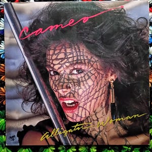May include: Album cover for Cameo's "Alligator Woman." The cover features a woman with black hair and face paint resembling a reptile pattern. The band's name is in pink script, and the album title is in yellow script.