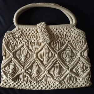 May include: A beige crocheted handbag with a knotted handle. The bag is made of a thick, rope-like material and has a delicate, intricate pattern.