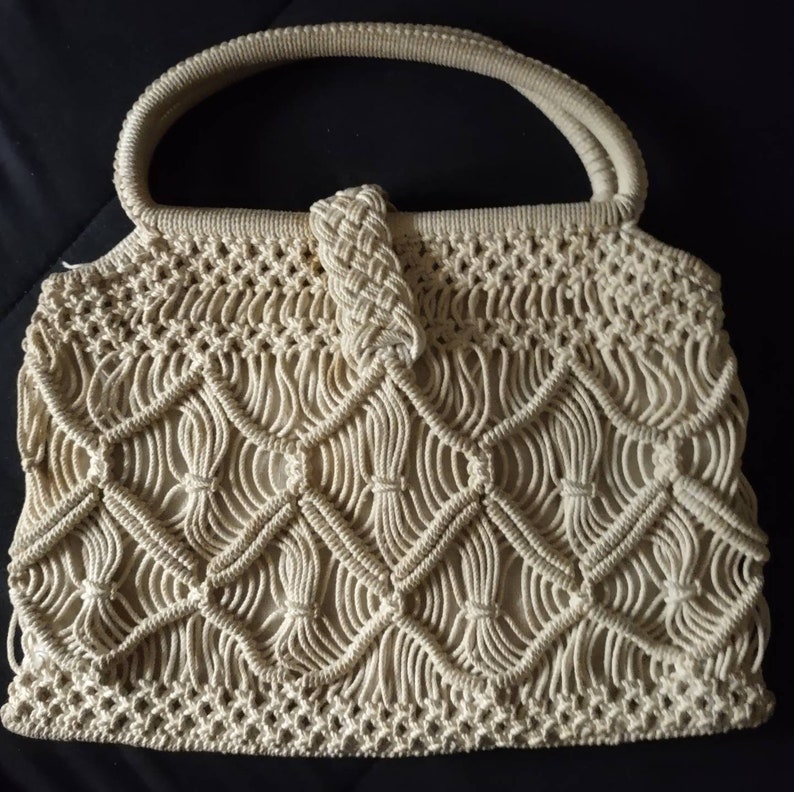 May include: A beige crocheted handbag with a knotted handle. The bag is made of a thick, rope-like material and has a delicate, intricate pattern.