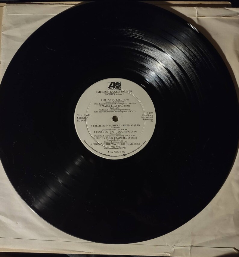 Puede incluir: Disco de vinilo negro con una etiqueta blanca. La etiqueta tiene el texto "Emerson Lake & Palmer Works Volume 2" y enumera los t&iacute;tulos de las canciones "So Far to Fall", "Maple Leaf Rag", "I Believe in Father Christmas", "Close But Not Touching", "Honky Tonk Train Blues" y "Show Me the Way to Go Home".