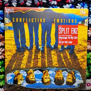 May include: A Split Enz vinyl record album titled "Conflicting Emotions." The album cover features a group of figures in blue pants and shoes, with their shadows cast on a yellow and brown background. A red sticker indicates the single "Message To My Girl."