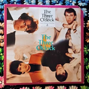 May include: A vintage album cover for "The Three O'Clock" featuring four band members. The album cover has a pink border and the band's name is in teal and white text. The album is titled "Active Without Traveling".