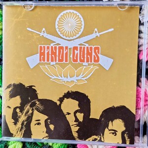 May include: A CD case with a gold cover featuring the band's name, "HINDI GUNS," in orange block letters. The cover art includes two crossed rifles, a circular emblem, and a lotus flower. Below the text are four band members.