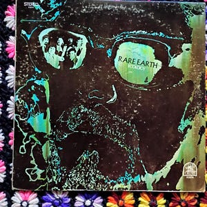 May include: Vintage vinyl record album cover for Rare Earth's "Ecology." The cover features a dark, abstract design with teal and green accents. The band's name and album title are visible. The album is placed on a colorful, crocheted blanket.