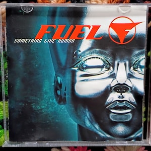 May include: A CD case featuring the album "Something Like Human" by Fuel. The cover art displays a metallic, futuristic human face in shades of blue and silver, with the band's name in red and a stylized "F" logo.